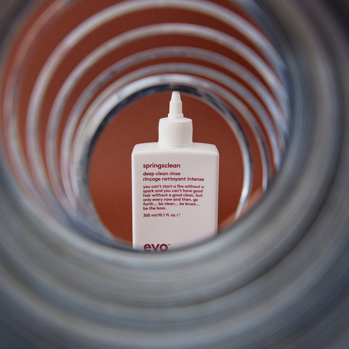 Eine weiße Flasche Springsclean Deep Clean Rinse von Evo ist durch eine gewundene Metallfeder vor einem braunen Hintergrund sichtbar.
