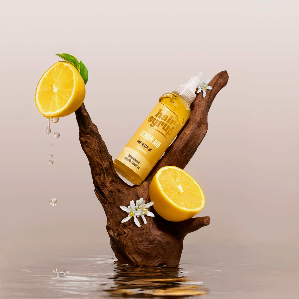 Eine Flasche Hair Syrup Lemon-Aid steht mit Zitronenhälften auf einem Holzast über dem Wasser, umgeben von Zitronentropfen und Blumen.