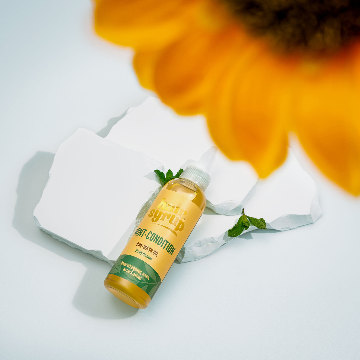 Eine Flasche Hair Syrup Mint Condition ruht auf weißen Steinen, begleitet von einer Sonnenblumenblüte und frischen Minzblättern.