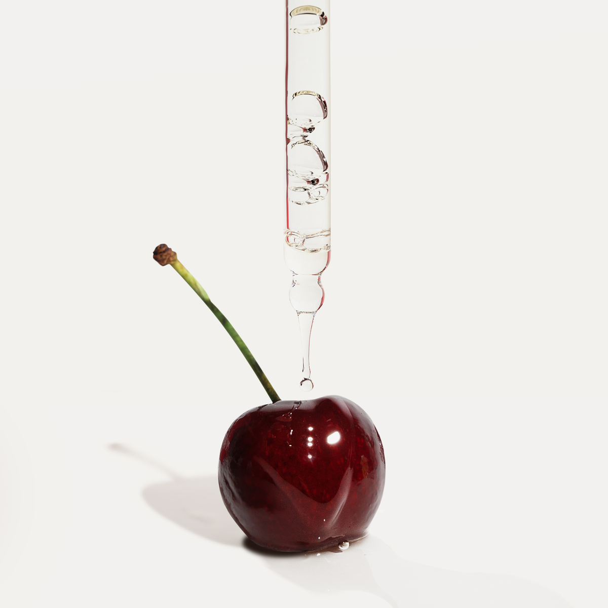 Eine Pipette verteilt die Flüssigkeit Very Cherry von Hair Syrup auf eine glänzende Kirsche mit langem Stiel vor weißem Hintergrund.