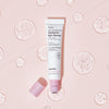 Offene Tube Hanskin Real Complexion Hyaluron Eye Serum mit rosa Deckel, neben klaren Geltropfen auf rosa Hintergrund.