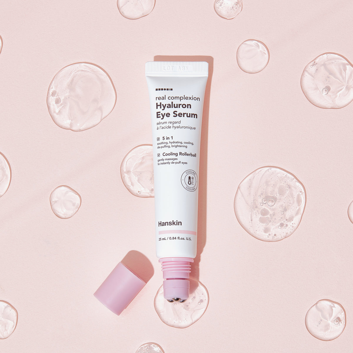 Offene Tube Hanskin Real Complexion Hyaluron Eye Serum mit rosa Deckel, neben klaren Geltropfen auf rosa Hintergrund.