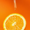 Eine Pipette gibt das Vitamin C Glow Serum auf eine frischen Orangenscheibe vor einem leuchtend orangefarbenen Hintergrund.