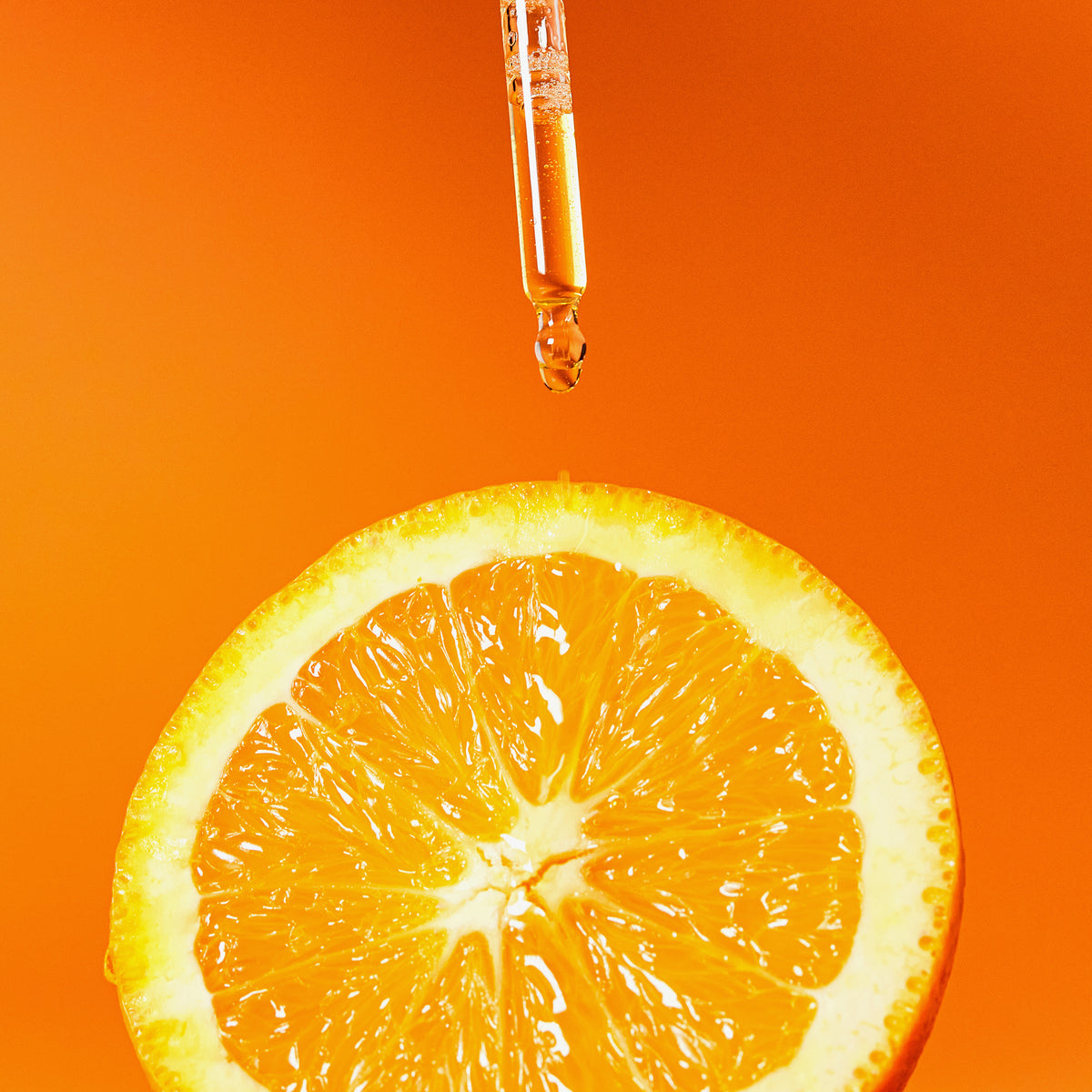 Eine Pipette gibt das Vitamin C Glow Serum auf eine frischen Orangenscheibe vor einem leuchtend orangefarbenen Hintergrund.