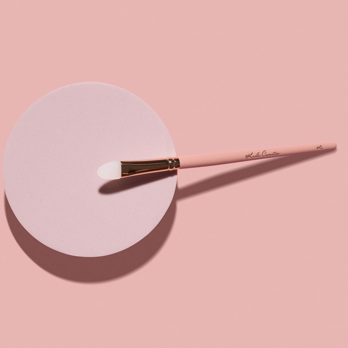 Der K1: Eye & Lip Glitter Pigment Brush in Pink liegt auf einem runden pinkfarbenen Zylinder, auf einem rosafarbenen Hintergrund.