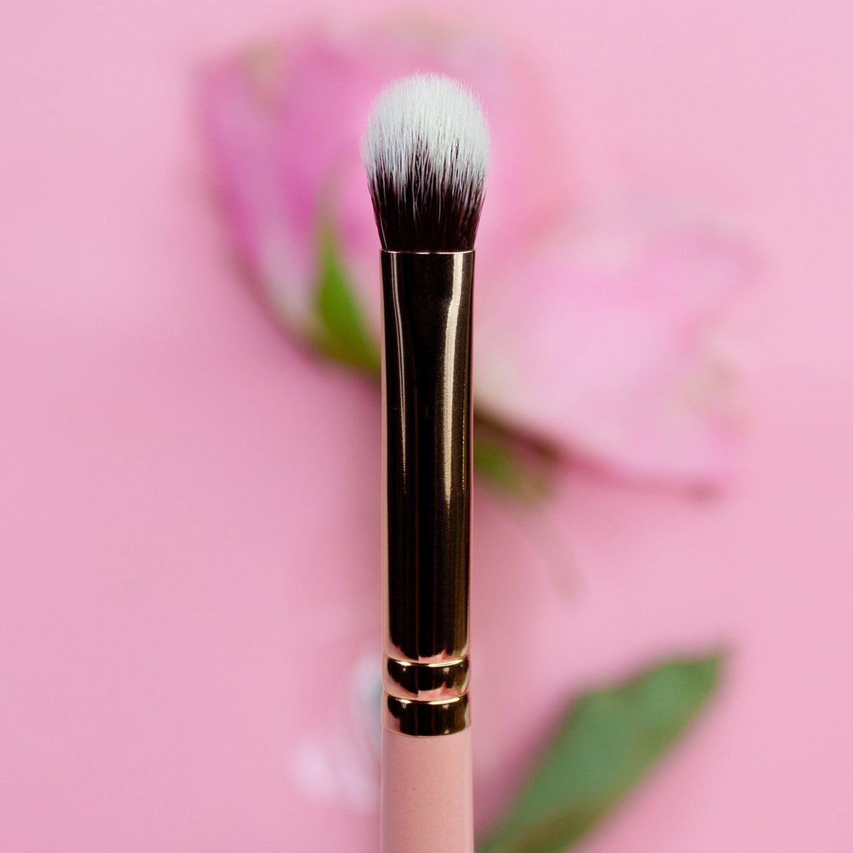 Der Make-up-Pinsel „K21:Pack and Blend“ mit goldenem Griff wird vor einem rosa Hintergrund mit einer verschwommenen Blume hervorgehoben.