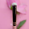 Nahaufnahme des Make-up-Pinsels K23: Short Shader mit rosa Griff vor einem rosa Hintergrund mit einer verschwommenen Blume.