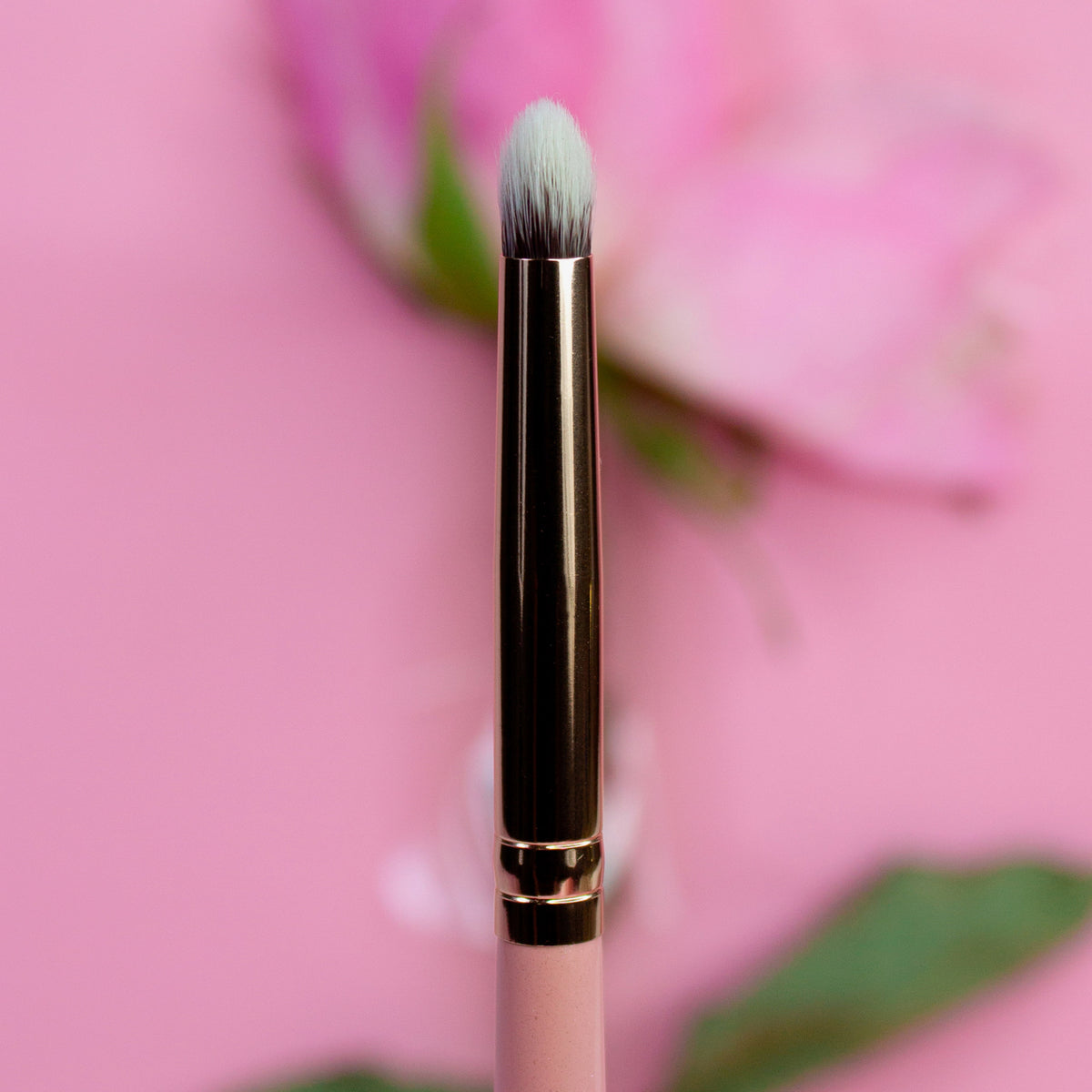 Der K24: Smoky Pencil, ein Make-up-Pinsel mit goldener Zwinge aufrecht vor einem rosa Hintergrund mit verschwommener Blume.