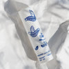 Eine weiße KORRES Lip Balm Tube mit blauen Vogelillustrationen liegt auf zerknülltem weißem Papier.