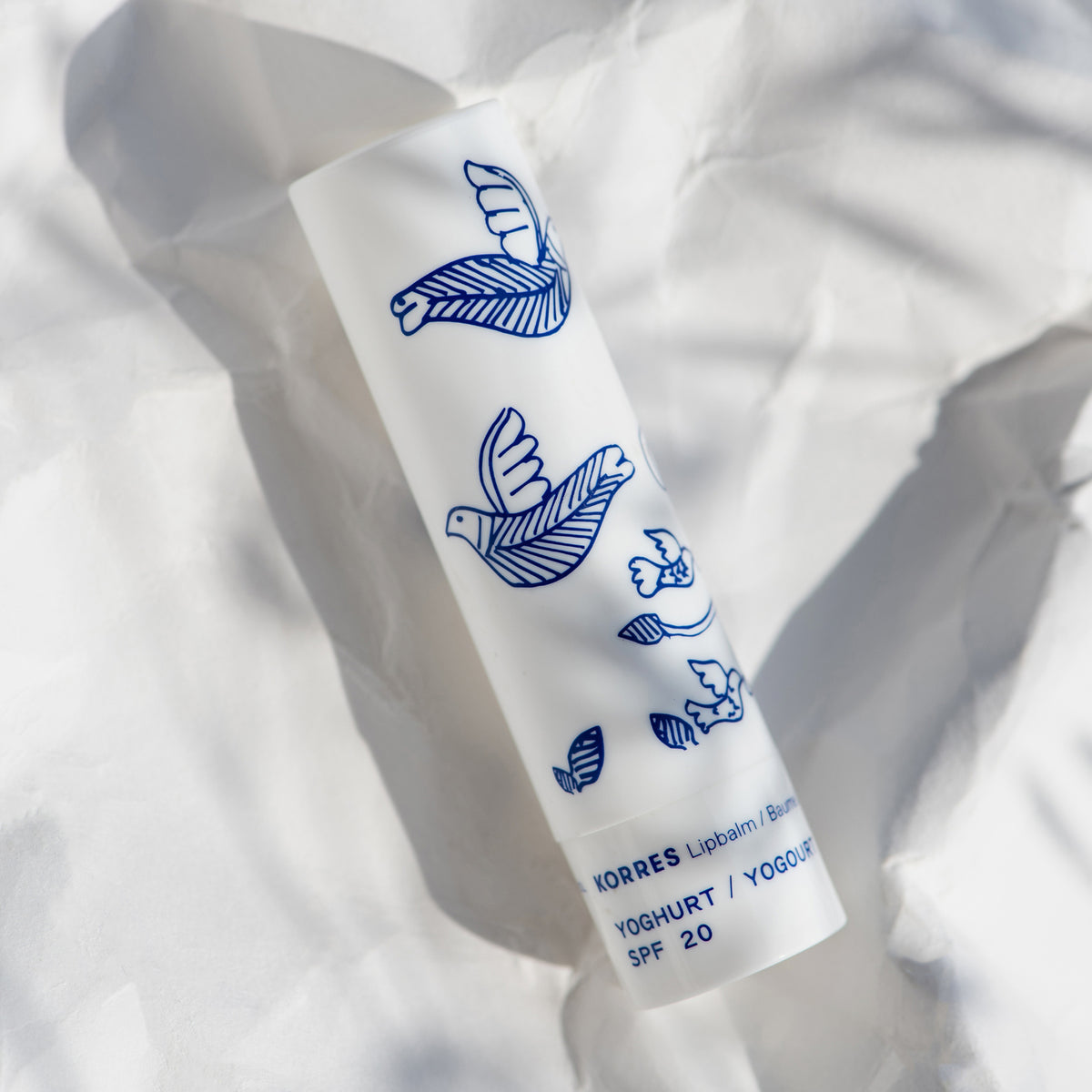 Eine weiße KORRES Lip Balm Tube mit blauen Vogelillustrationen liegt auf zerknülltem weißem Papier.
