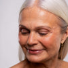 Eine ältere Frau mit langem grauem Haar und geschlossenen Augen lächelt sanft, präsentiert Artistick Blush Muse von LH Cosmetics vor einem hellen Hintergrund.