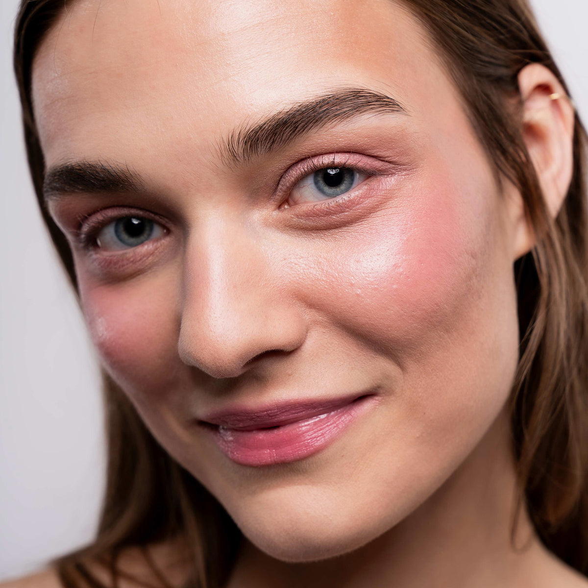 Eine Person mit heller Haut, blauen Augen und dem rosafarbenen Artistick Blush von LH Cosmetics lächelt leicht in die Kamera.