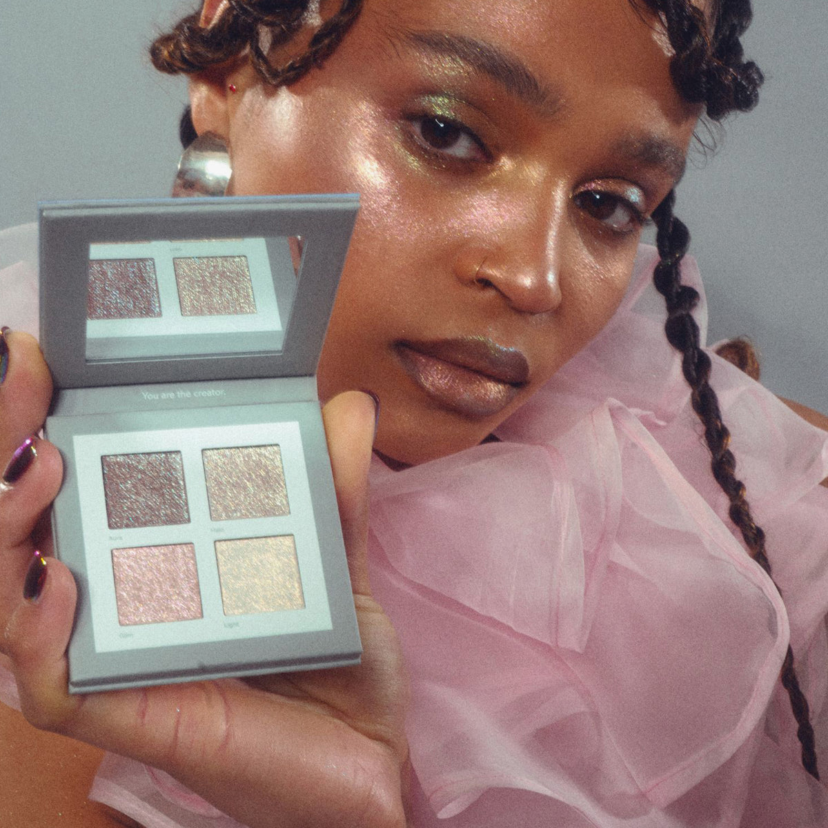Eine Person in rosafarbenem, durchsichtigem Stoff hält die Aurora-Palette von LH Cosmetics mit vier schimmernden Lidschatten in der Nähe ihres glitzernden Gesichts.