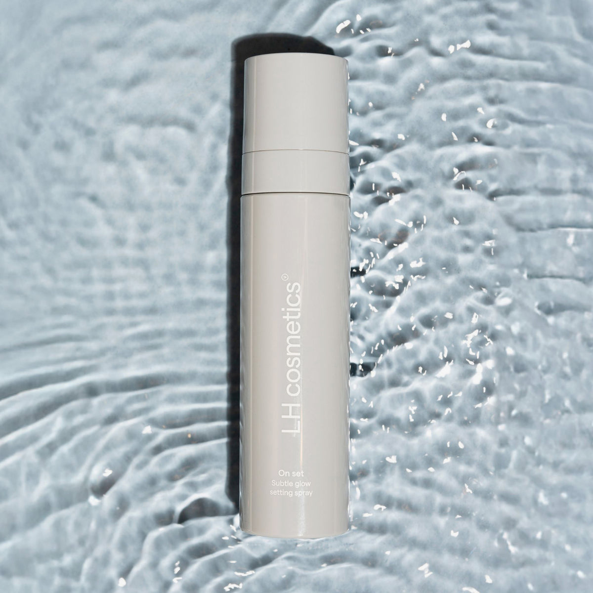 Eine Flasche LH Cosmetics On Set – Subtle Glow Setting Spray steht auf plätscherndem, reflektierendem Wasser.