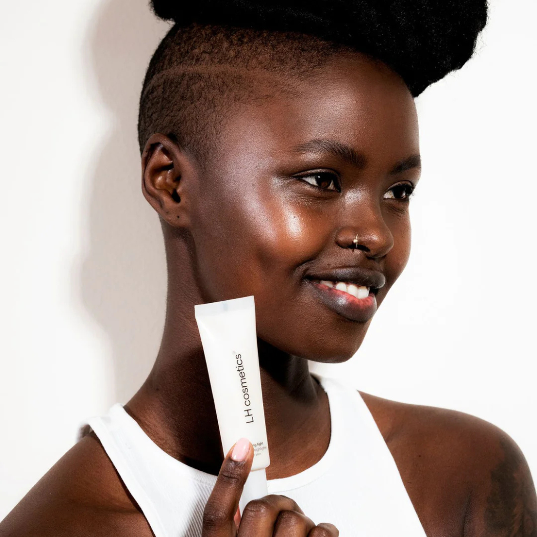 Eine lächelnde Person mit strahlenderm dunklen Teint hält eine Tube Shaping Light Cool Glow von LH Cosmetics.