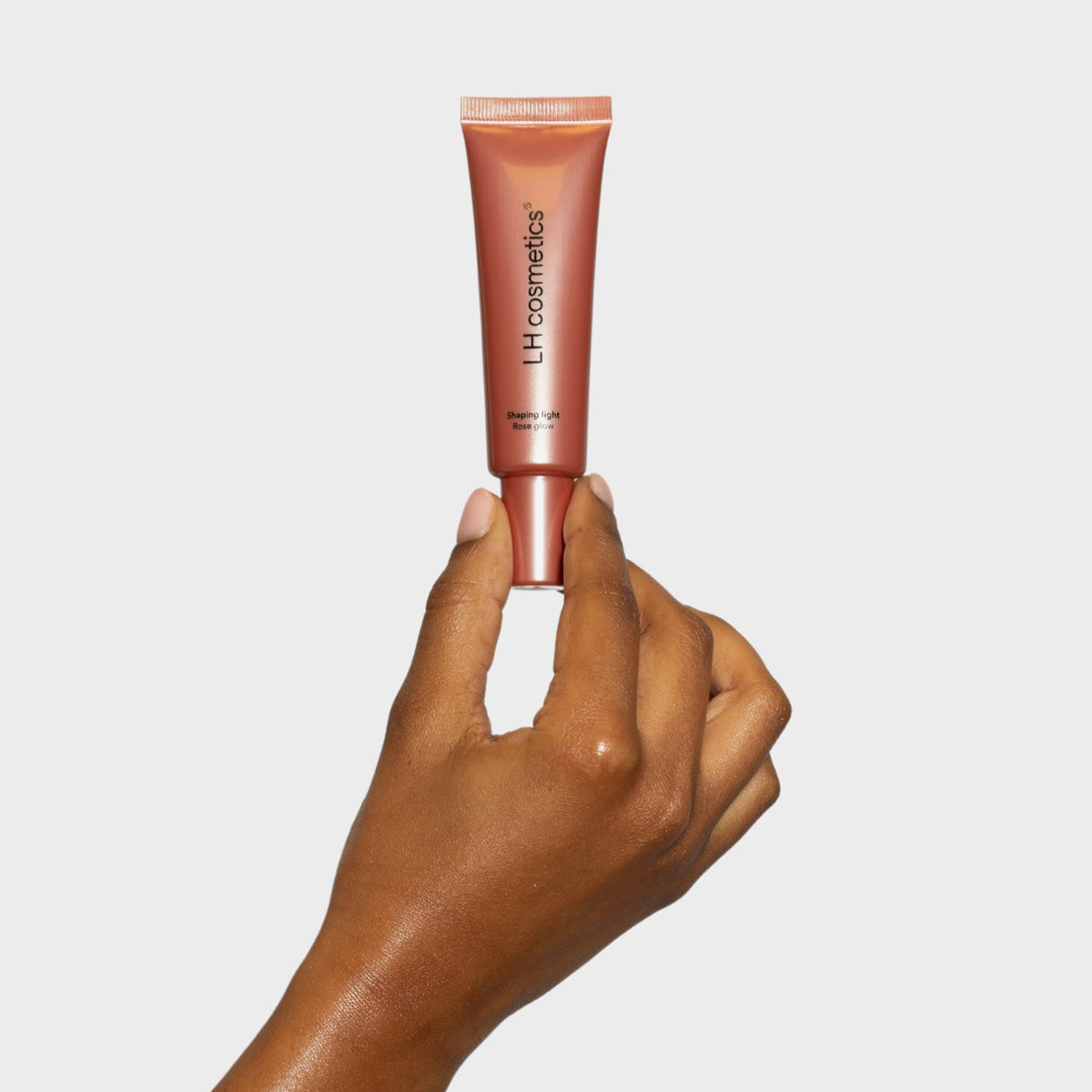 Eine Hand hält eine roséfarbene Tube LH Cosmetics Shaping Light Rose Glow vor schlichtem weißen Hintergrund.