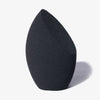 The Other Sponge von LH Cosmetics, ein schwarzer Make-up-Schwamm in Tropfenform mit schräger Kante, steht aufrecht auf einem hellgrauen Hintergrund.