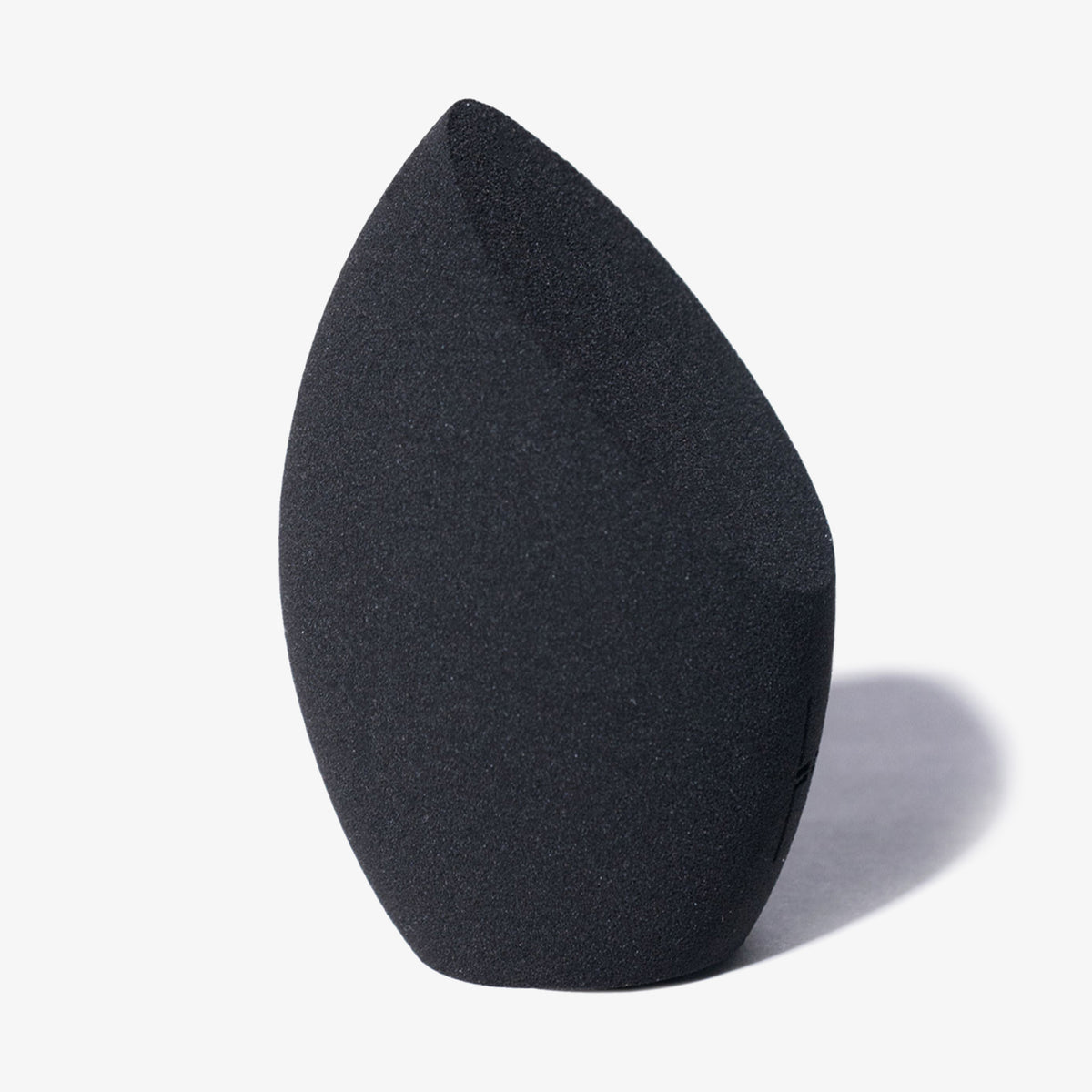 The Other Sponge von LH Cosmetics, ein schwarzer Make-up-Schwamm in Tropfenform mit schräger Kante, steht aufrecht auf einem hellgrauen Hintergrund.