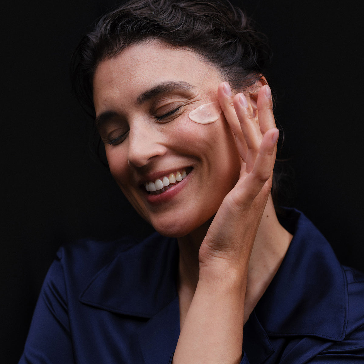 Eine Frau lächelt mit geschlossenen Augen und trägt vor einem dunklen Hintergrund Lumene NORDIC BLOOM [LUMO] Anti-Falten & Straffe Tagescreme auf ihre Wange auf.