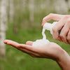 Eine Person verteilt weißen Schaum 
 des Lumene NORDIC HYDRA [LAHDE] Arctic Aqua Foaming Cleanser auf ihrer Handfläche im Freien, mit Pflanzen im Hintergrund.