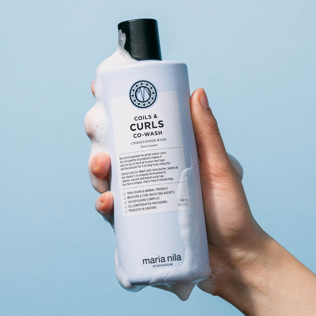Eine Hand hält eine schaumige Flasche der maria nila Coils & Curls Co-Wash Spülung vor einem blauen Hintergrund.