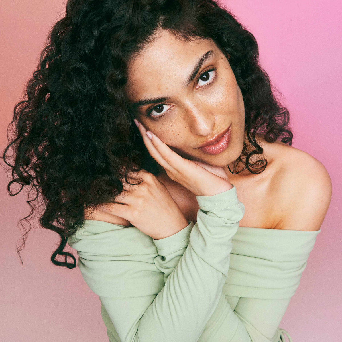 Eine Frau mit lockigem schwazrem Haar und Sommersprossen posiert vor einem rosafarbenen Hintergrund und präsentiert ihre mit der maria nila Coils & Curls Finishing Treatment Masque gestylten Locken.