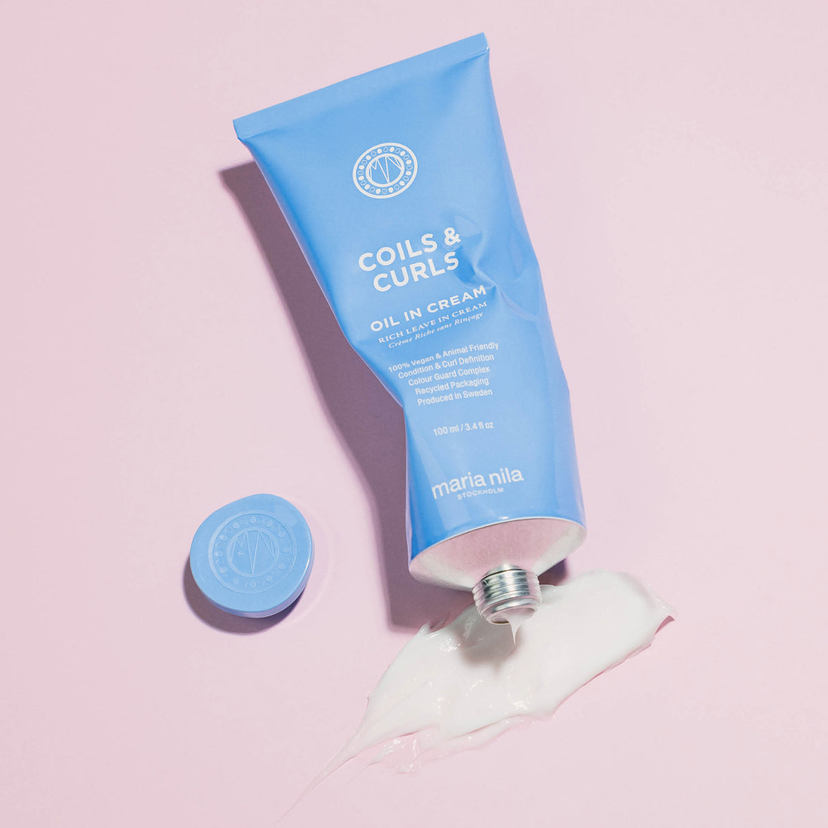 Blaue geöffnete Tube maria nila Coils & Curls Oil-In-Cream ist auf einem rosafarbenen Hintergrund abgebildet, daneben ist etwas Creme ausgepresst.