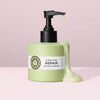 Eine grüne Pumpflasche mit der maria nila Structure Repair Leave-in Cream, bei der die Creme an der Seite herunterläuft, auf einem rosa Hintergrund.