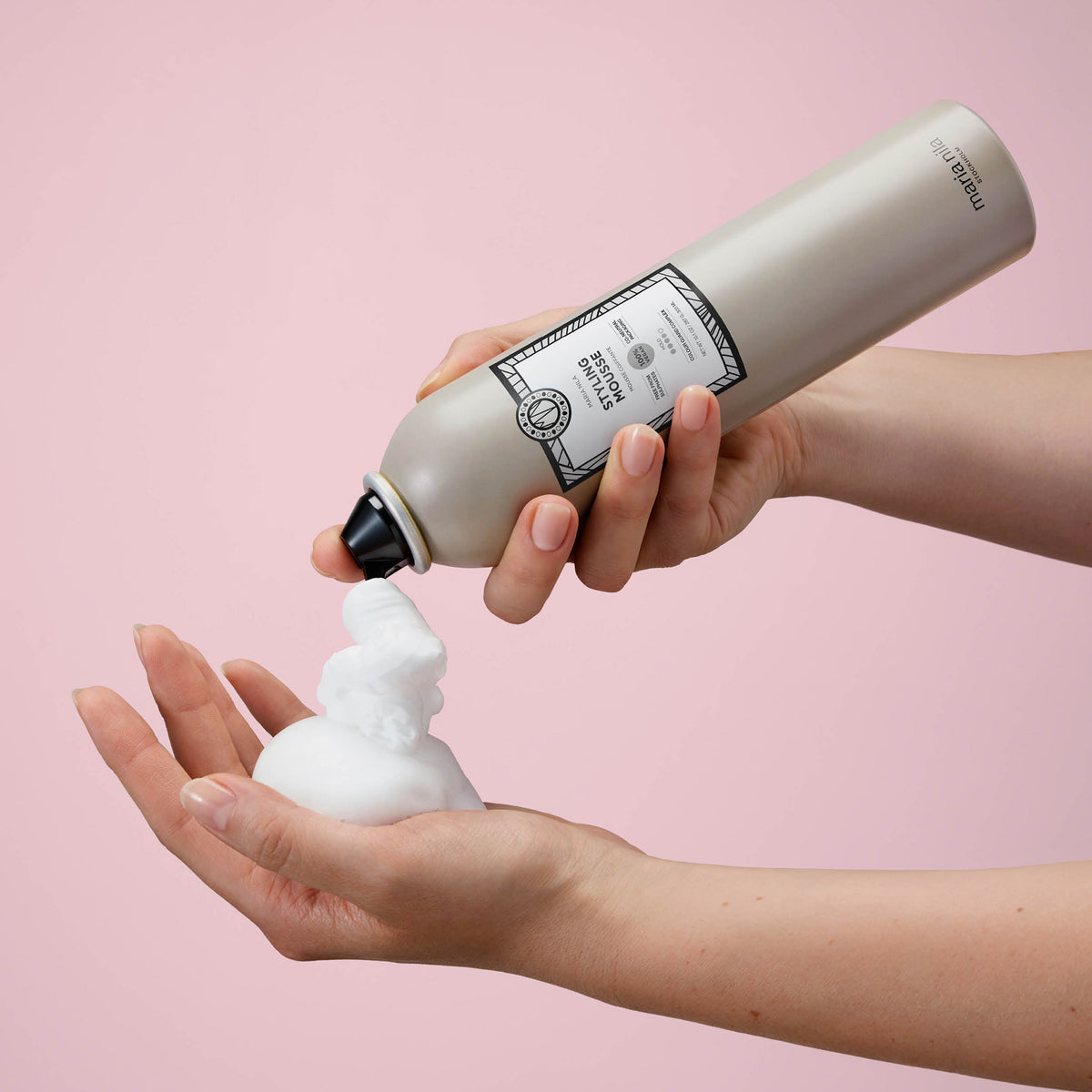 Eine Person gibt weißes maria nila Styling Mousse vor einem rosafarbenen Hintergrund in ihre Hand.