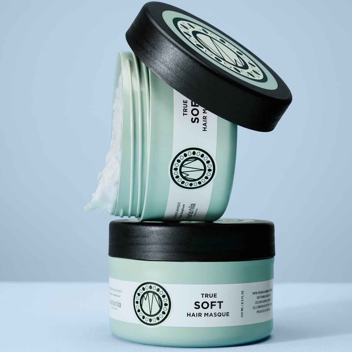 Zwei mintgrüne True Soft Masque-Behälter von maria nila sind auf einem hellblauen Hintergrund gestapelt, wobei einer geöffnet ist.