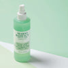 Eine Flasche Mario Badescu Gesichtsspray mit Aloe, Gurke und grünem Tee ist auf einem pastellfarbenen Hintergrund abgebildet.