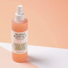 Eine Flasche Gesichtsspray mit Aloe, Kräutern und Rosenwasser ist vor einem pfirsichfarbenen und weißen Hintergrund abgebildet.
