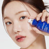 Eine Frau trägt die medicube One Day Exosome Shot Pore Ampoule 2000 aus einer blauen Flasche auf ihre Wange auf.
