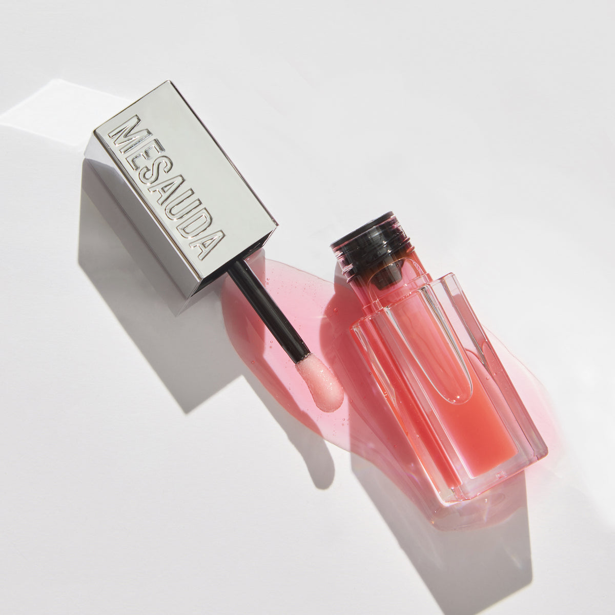 Eine geöffnete Tube des rosafarbenen Lipglosses LIPOILOGY™ von Mesauda Beauty mit einer silbernen Kappe ist auf einer weißen Fläche verschüttet.