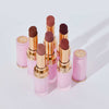 Sechs geöffnete Beyond Blurry-Lippenstifte von NABLA in verschiedenen Nude- und Rosétönen stehen aufrecht nebeneinander. Die rosafarben-goldenen Lippenstifthülsen glänzen im Licht, zwei passende Kappen liegen daneben.
