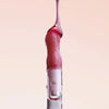 Ein rötlicher Tropfen Lip Candy Oil Black Cherry gleitet vor einem hellrosa Hintergrund über den Applikator. 