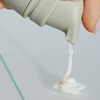 Eine Hand drückt das NERDS. 10% Azelainsäure Cremeserum aus einer grauen Tube auf eine klare Glasfläche.
