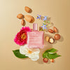 Eine Flasche NUXE Huile Prodigieuse® Or Florale ist von Blumen, grünen Blättern, Nüssen und Samen auf einem beigen Hintergrund umgeben.