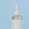 Nahaufnahme des Round Lab 1025 Dokdo Cleansing Oil mit Pumpspender, aus dem ein einzelner Tropfen Öl herabrinnt, vor blauem Hintergrund.