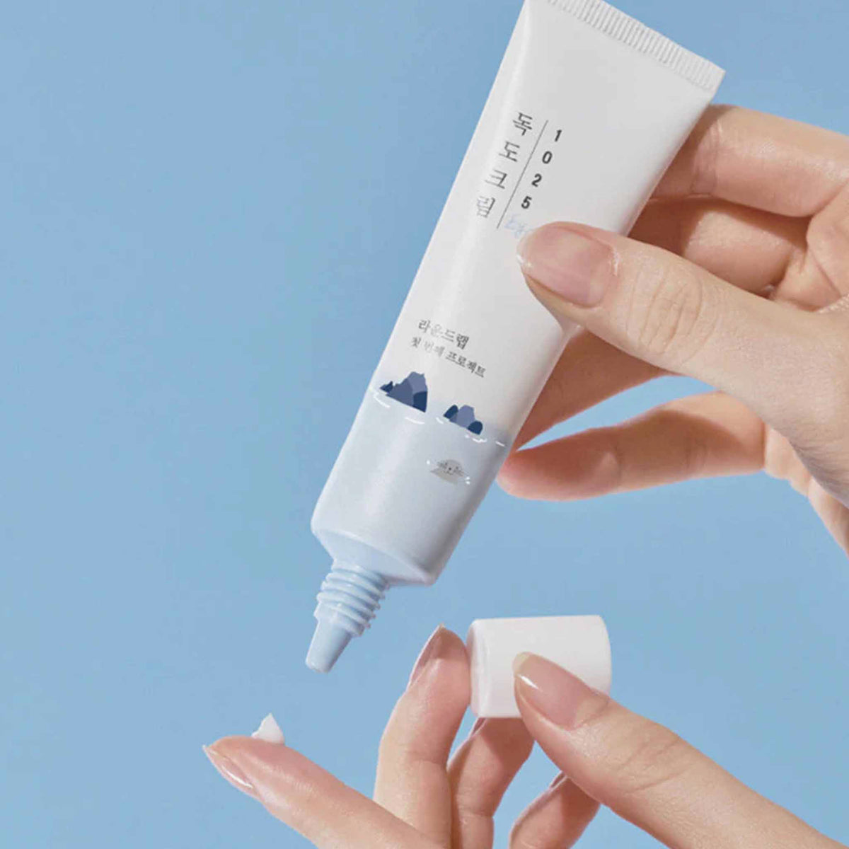 Runde Tube der Round Lab 1025 Dokdo Eye Cream mit hellblauem Design wird auf einen Finger aufgetragen.