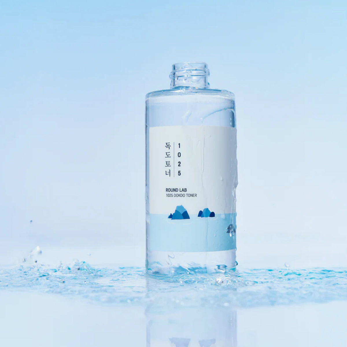 Eine durchsichtige Flasche ROUND LAB 1025 Dokdo Toner steht auf einer nassen Oberfläche vor einem blauen Hintergrund.