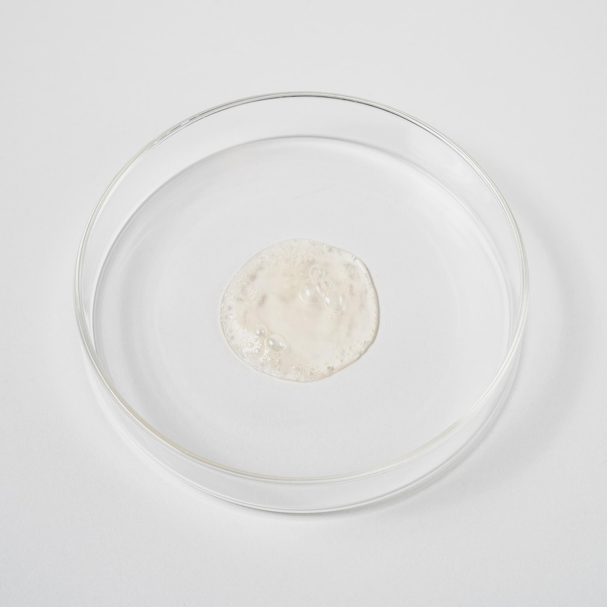 Eine durchsichtige Petrischale enthält in der Mitte eine kleine Menge des durchsichtigen, sprudelnden Bio-Pilixin® Shampoo+Women von Scandinavian Biolabs auf weißem Hintergrund.