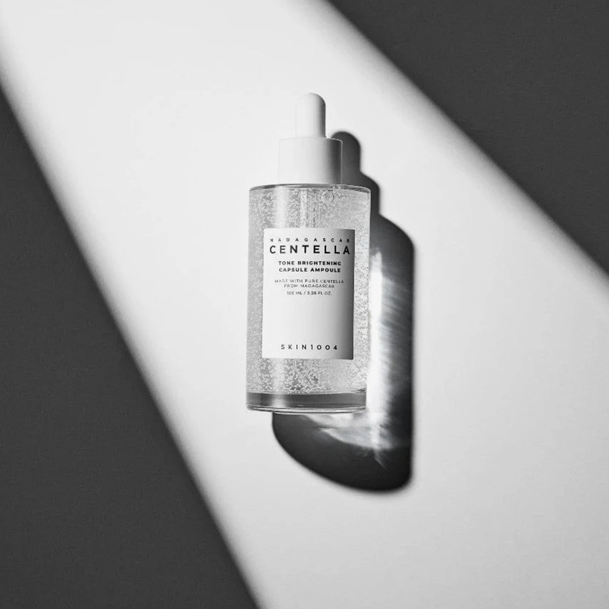 Eine Serum-Tropfflasche mit der Aufschrift „Madagascar Centella Tone Brightening Capsule Ampoule“ steht auf einem weiß-grauen Hintergrund mit Schatten.