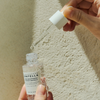 Eine Hand hält eine Pipette über einer offenen Flasche Madagascar Centella Tone Brightening Capsule Ampoule vor einer beige strukturierten Wand.