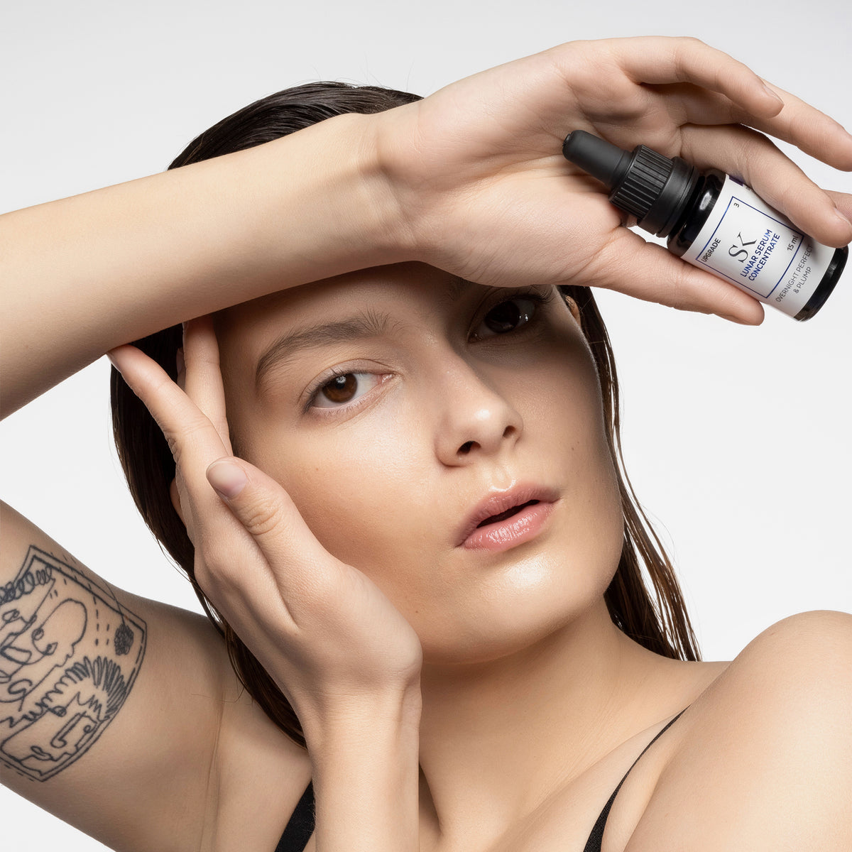 Eine Frau mit einer Tätowierung auf dem Arm hält eine Tropfflasche Lunar Serum in die Nähe ihres Gesichts und behält dabei einen neutralen Gesichtsausdruck bei.