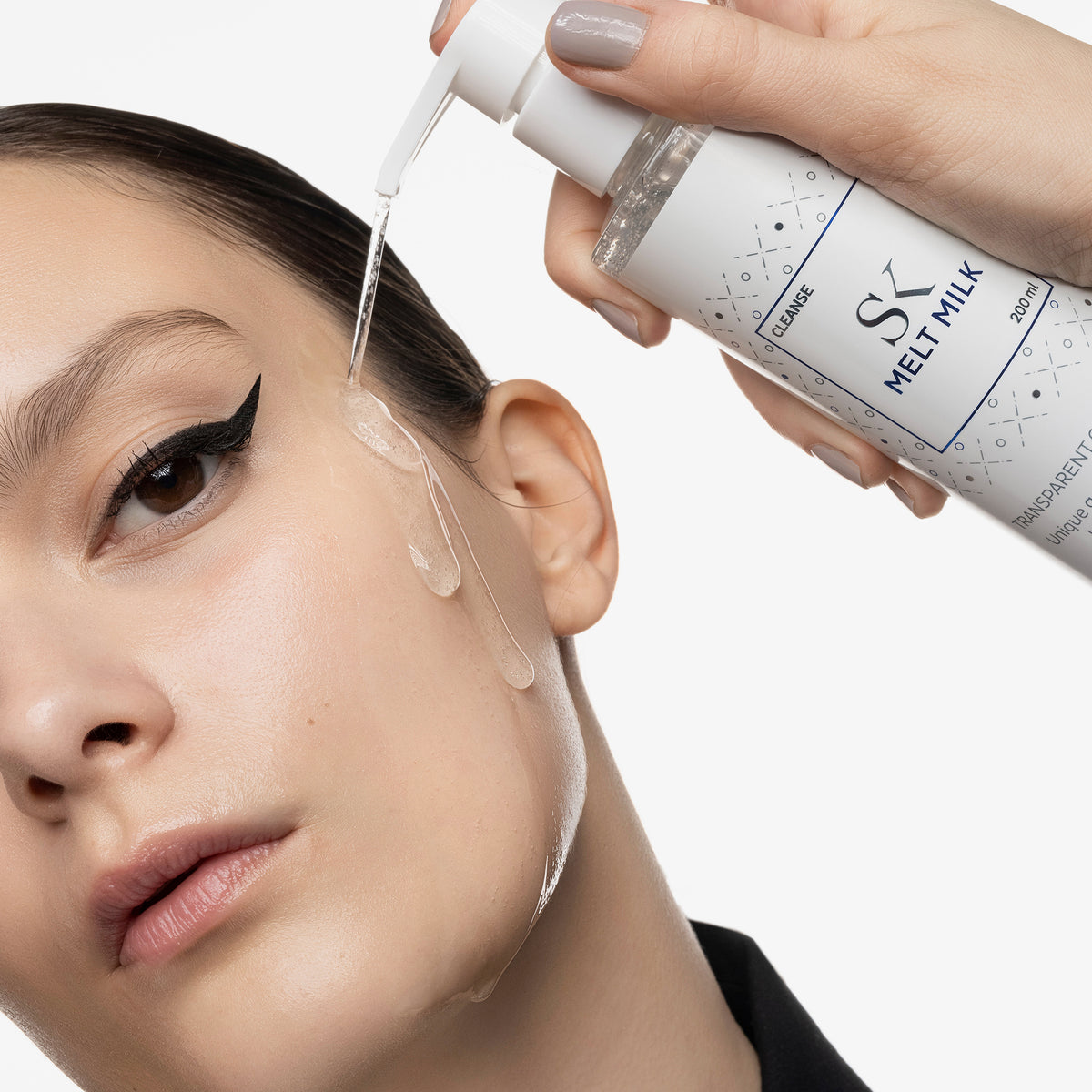 Nahaufnahme einer Frau mit Eyeliner, auf deren Gesicht eine klare Reinigungsmilch aus einer Pumpflasche aufgetragen wird.