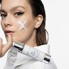 Frau mit weißem X aus Creme auf der Wange hält die SK O/P Hydra Cream in der Hand, trägt Eyeliner und blickt direkt in die Kamera vor hellem Hintergrund.
