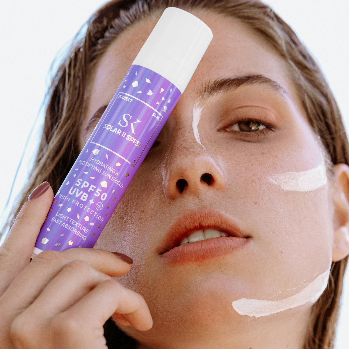 Eine Frau hält eine Flasche Solar II SPF50-Sonnenschutzmittel in der Hand vor ihr Gesicht; das Produkt ist auf ihr Gesicht aufgetragen.