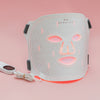 Weiße STYLPRO LED-Gesichtsmaske mit roten Lichtimpulsen, Kabel und Steuergerät, auf rosa Hintergrund stehend und startbereit.