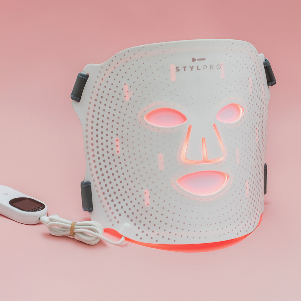 Weiße STYLPRO LED-Gesichtsmaske mit roten Lichtimpulsen, Kabel und Steuergerät, auf rosa Hintergrund stehend und startbereit.
